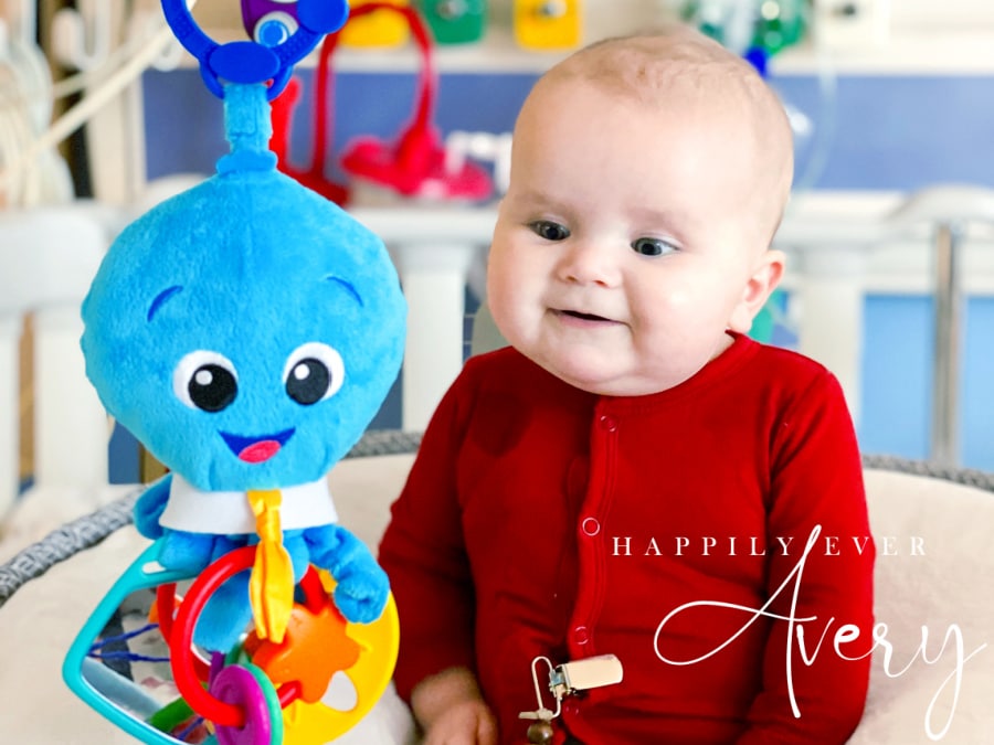 infant in red sleeper smiling with blue octopus toy hanging beside her