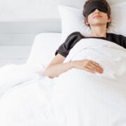 woman lying with a sleeping mask in bed under white comforter blanket