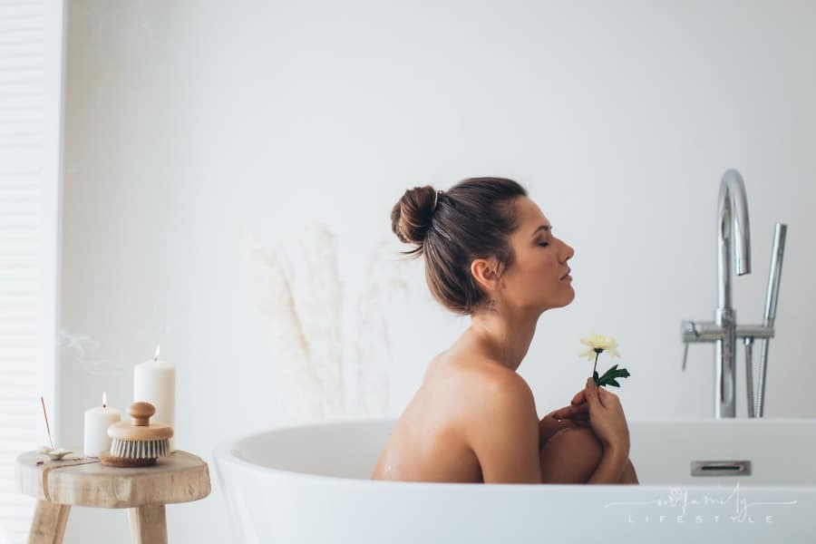 woman sitting up in a bathtub holding a yellow flower with table of candles nearby