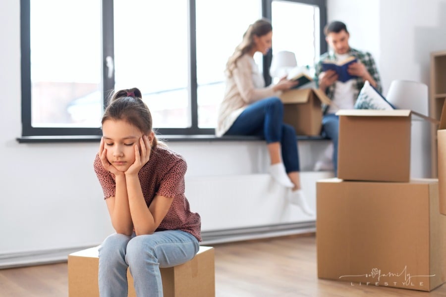 sad girl sitting on moving box sad about moving to new home while parents talk behind her