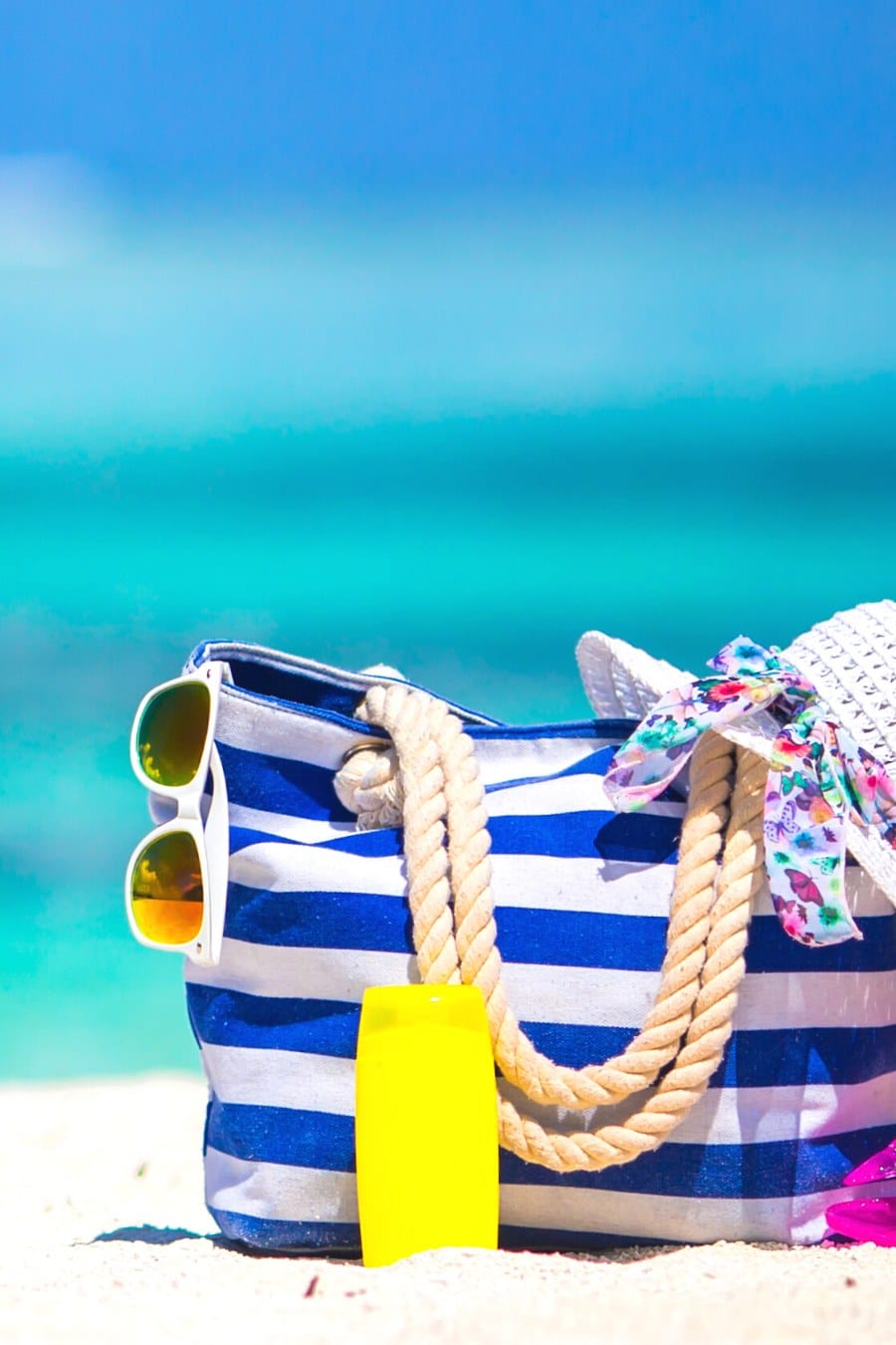 blue and white striped beach bag with sunglasses, sunblock and a hat sitting on sandy beach