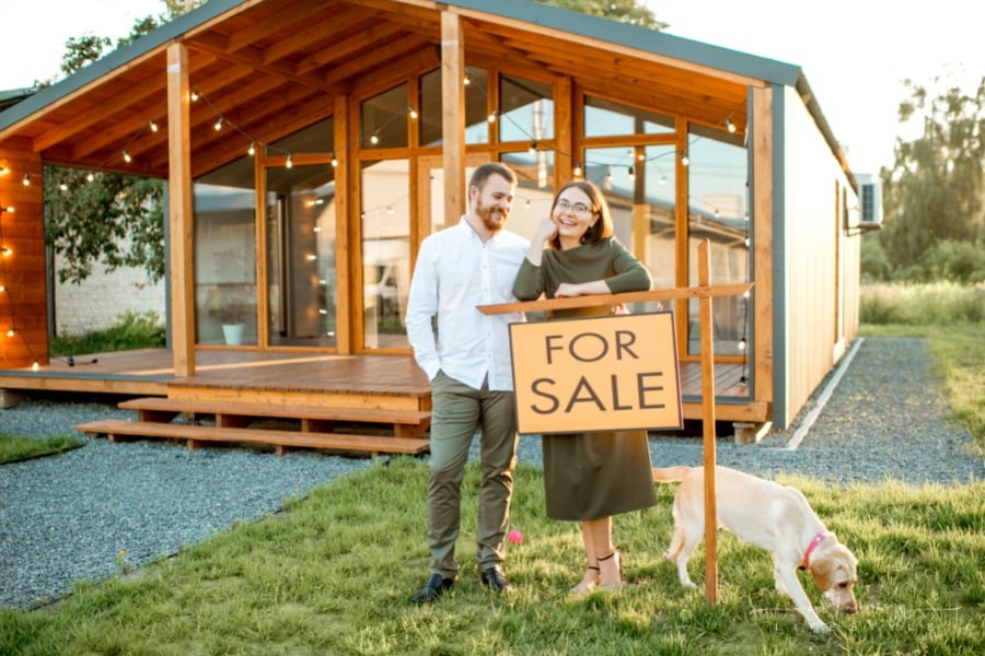 couple standing with dog in front of wooden country house with for sale sign in yard