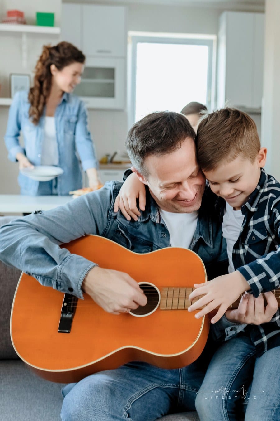 father and son playing guitar together while mom and second son set kitchen table in background