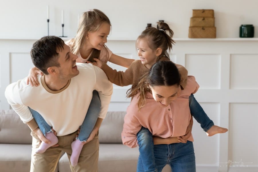 preschooler daughters piggyback with young parents as they play together in living room at home, loving mom and dad carry small daughters on back while having fun engaged in fun activity