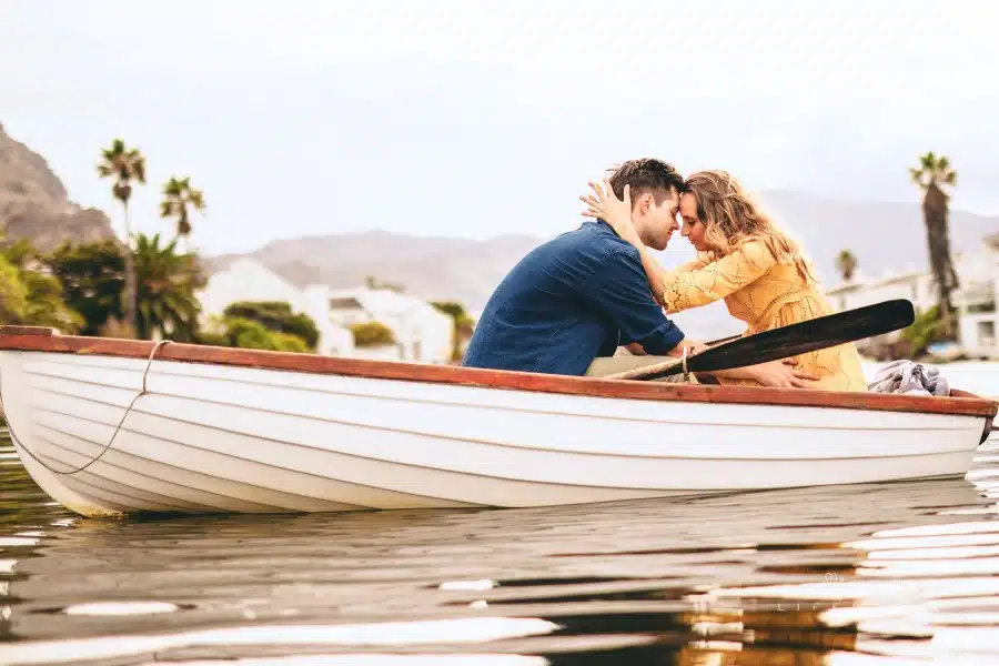 Young couple sitting together in a boat touching their heads. Couple in love on a boat date in a lake with hills and houses in the background.