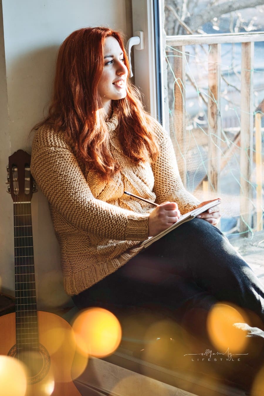 woman with red hair sitting in window, looking outside and writing in notebook with guitar nearby