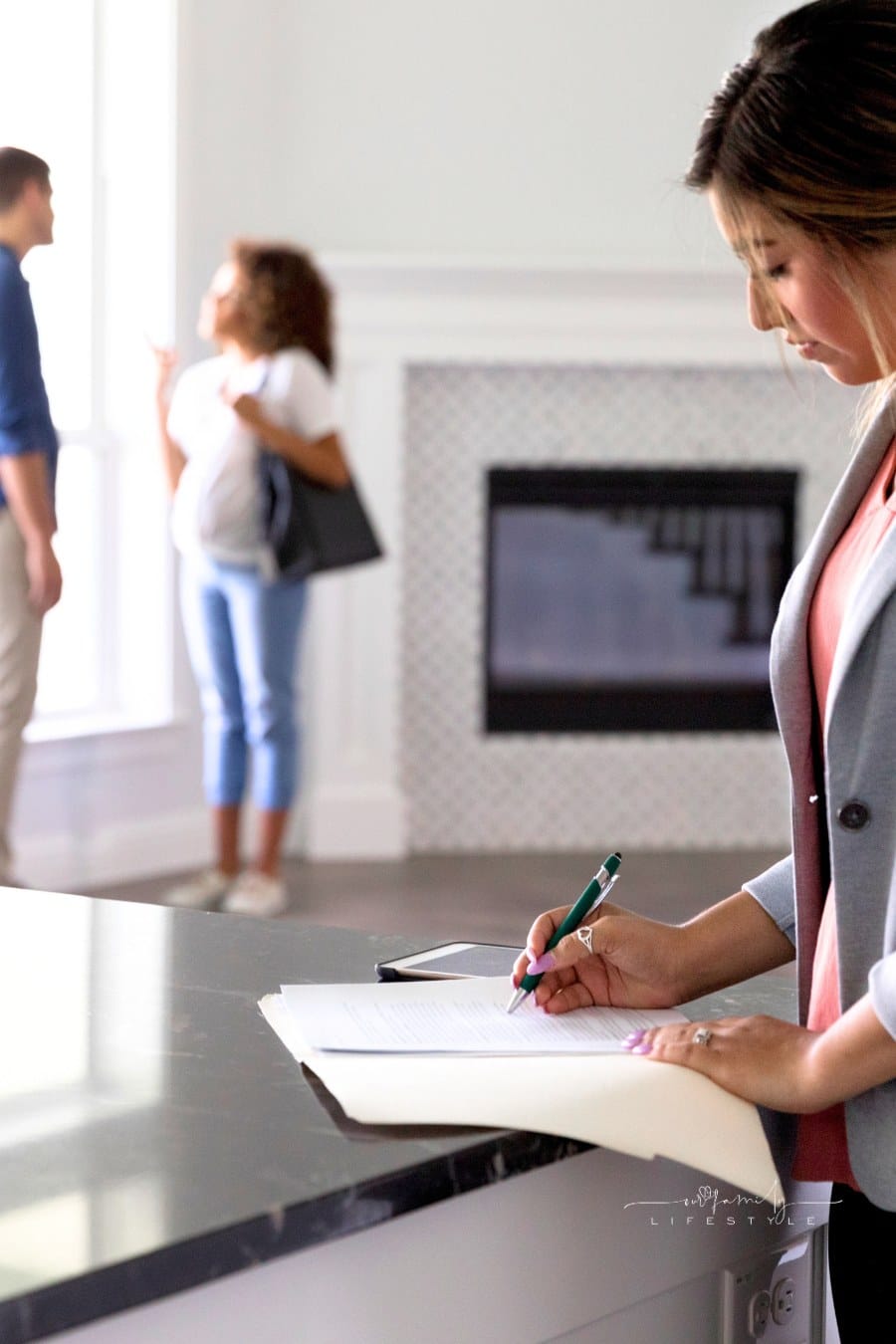 As a young couple have a discussion about buying a new home, a female realtor is in the foreground reviewing documents.