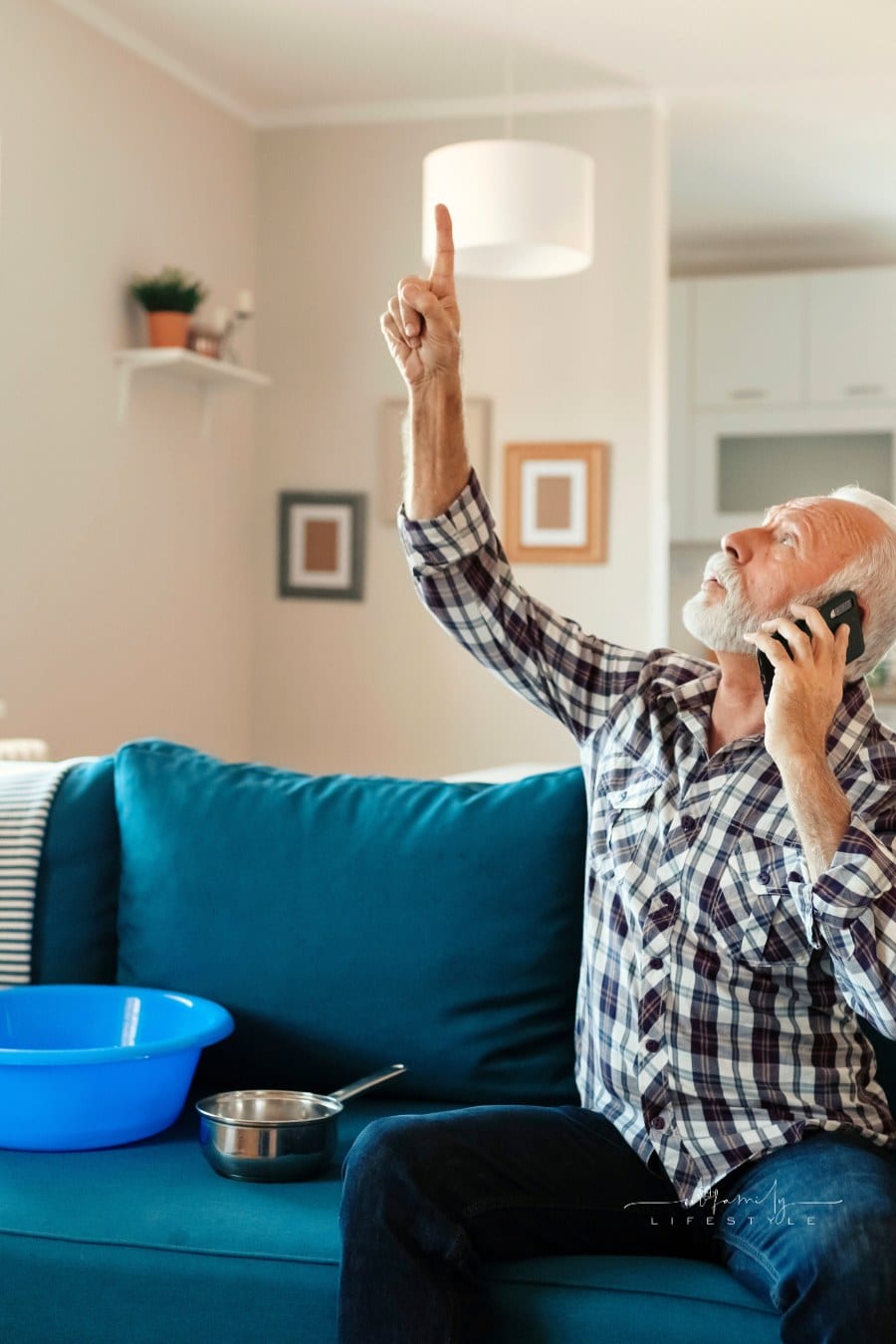 Angry Bearded Senior Man Talking on Smartphone Near Plastic Wash Bowl at Home in the Living Room Because of Roof Leaking
