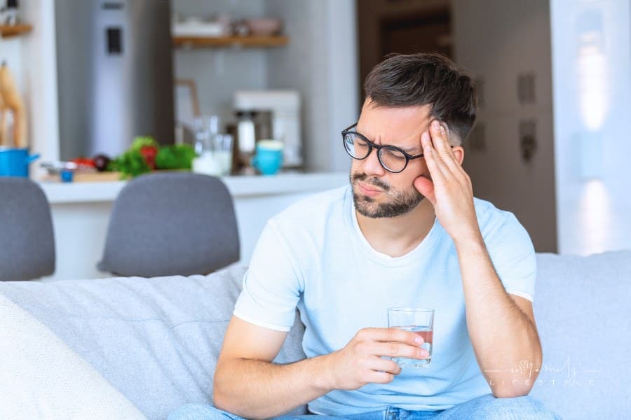 young man suffering from headache while sitting on couch with a glass of water